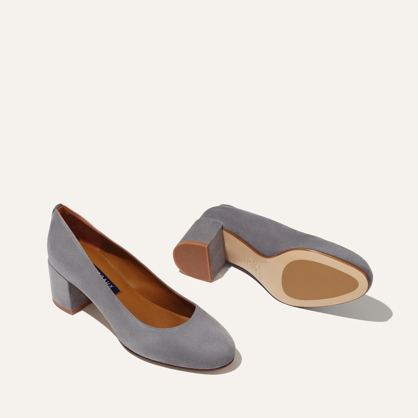 Margaux’s The Heel in Steel Suede—one elegant block-heeled pump stands upright to show the top and side, while the other lies sideways revealing sole and heel, both displayed against a plain off-white background.