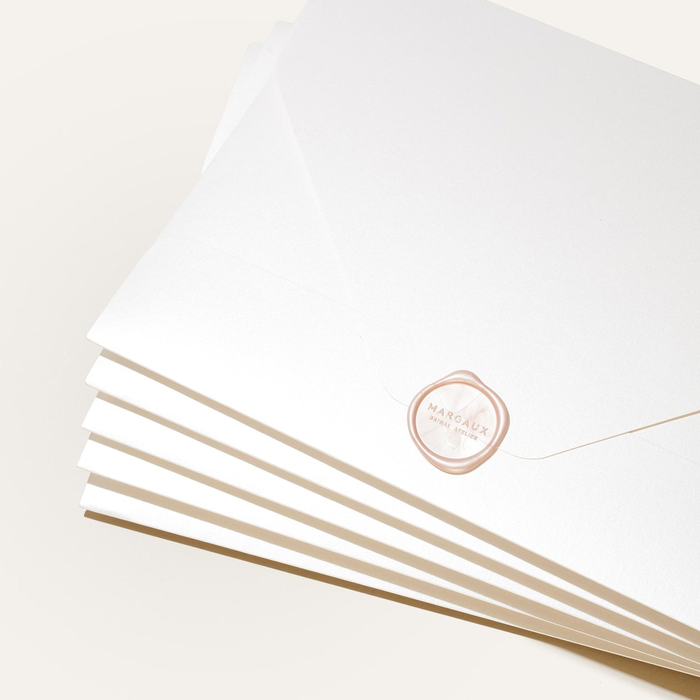 A stack of white envelopes, each sealed with a pale pink wax seal stamped “MARGAUX” in gold, displays the Margaux Bridal Swatch Kit—an elegant introduction to the brand’s bespoke bridal collection. The envelopes are fanned out on a neutral background.