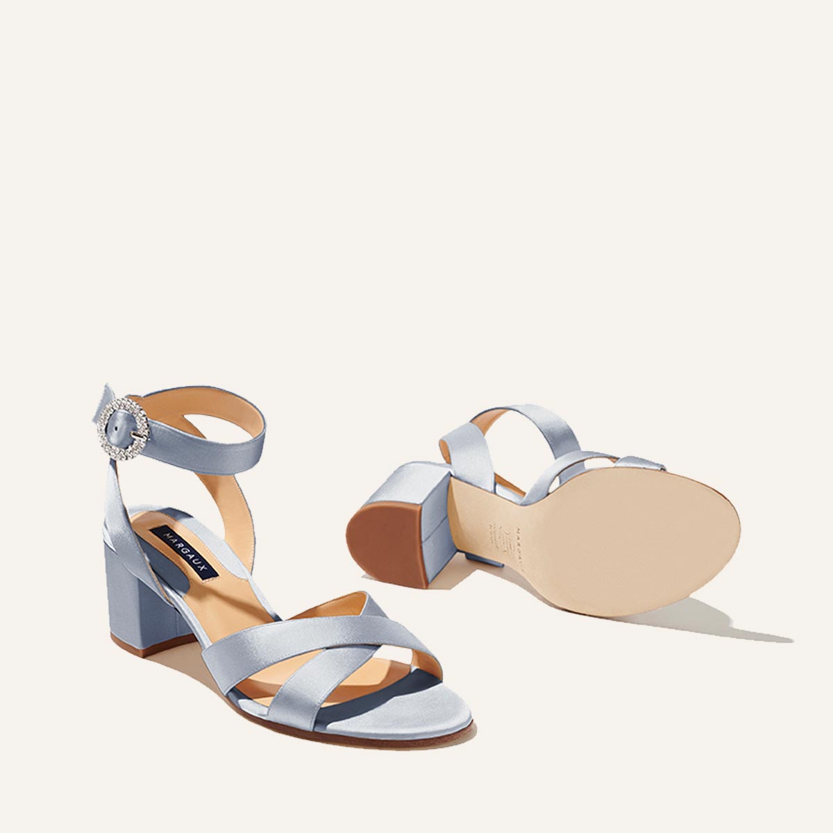 The City Sandal by margaux-development features light blue powder satin, open toes, crisscross and ankle straps with a crystal buckle, and a cushioned insole for comfort. One sandal stands upright; the other lies on its side to display the details.