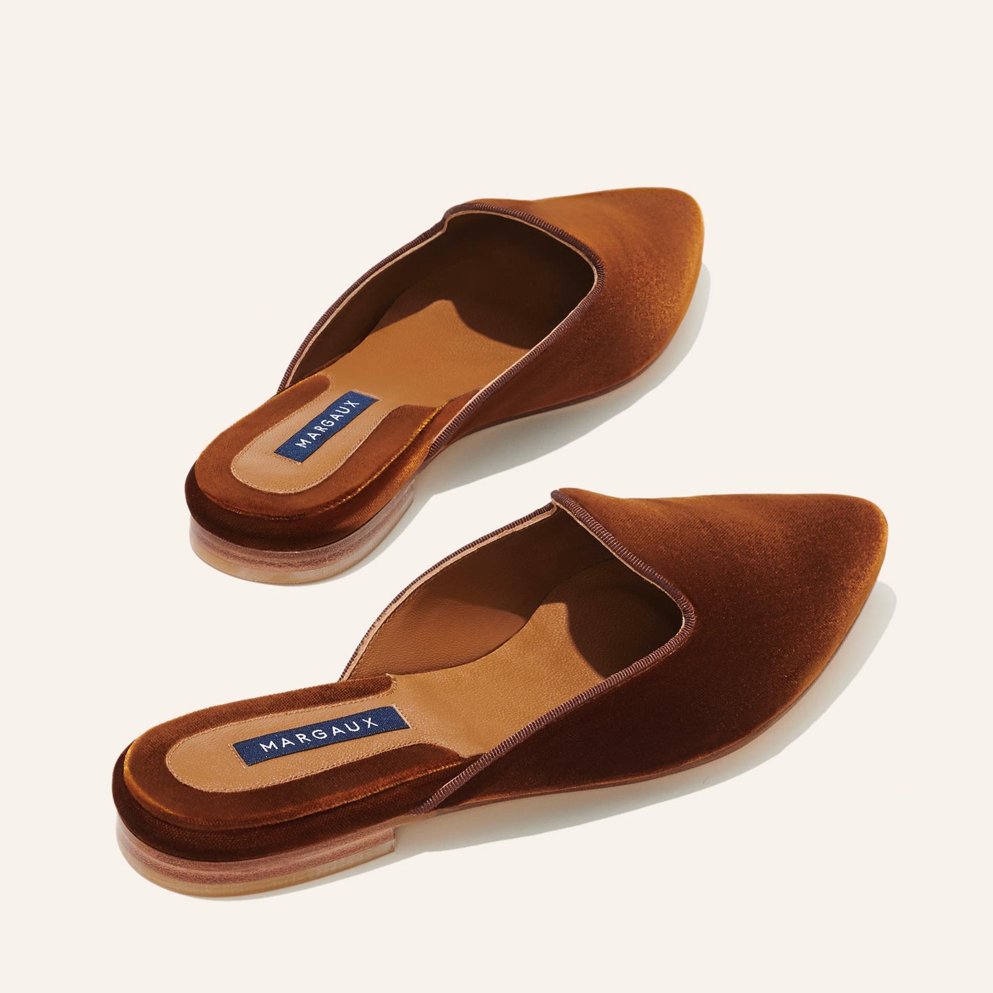 The Mule - Rust Velvet by Margaux features brown velvet pointed-toe slides with tan insoles and a branded footbed, displayed on a light background. These elegant mules offer effortless style and comfort.