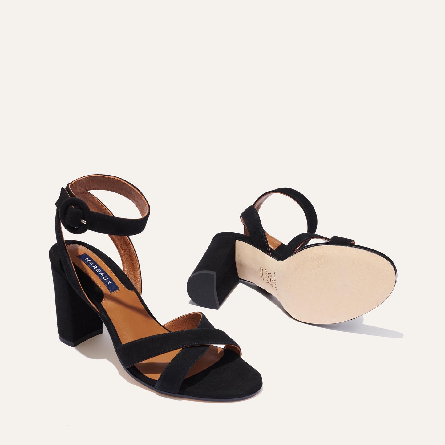 The Uptown Sandal - Black Suede by margaux-development features block heels, ankle straps with buckles, and a comfortable sole—one shoe stands upright while the other lies on its side, highlighting its design for all-day wear.