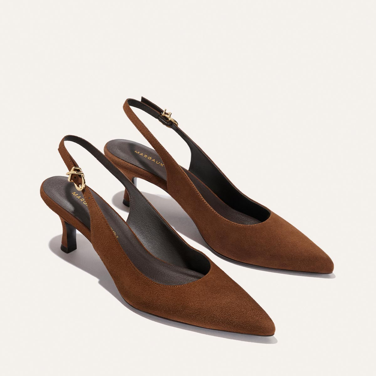 A pair of The Colette - Ganache Suede heels by Margaux, featuring brown suede, snip nose toes, micro heels, and gold buckle ankle straps, displayed on a light background.
