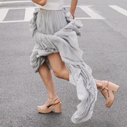 Wearing The Bardi - Petal Calf heels by margaux-development, a person strides briskly down the street in a flowing, light gray pleated skirt, the fabric billowing with their movement.
