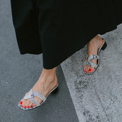 A person wearing a long black skirt and open-toed margaux-development The MX 35 - Natural Python Embossed sandals with block heels and red nail polish stands elevated on the edge of a street.