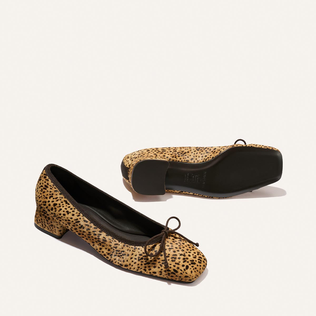 Margaux’s The Ada in Tan Mini Cheetah Haircalf: ballet flats featuring a tan cheetah print, small bow detail, square toe, and low block heel. One shoe is shown from the side; the other displays its black sole.