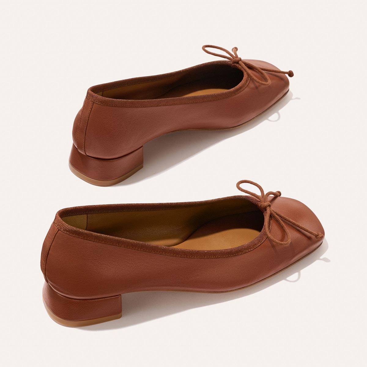 The Ada - Cognac Nappa by margaux-development: brown leather ballet pumps with low block heels, rounded toes, sacchetto construction for a glove-like fit, and thin bow ties on the front, shown on a light background.