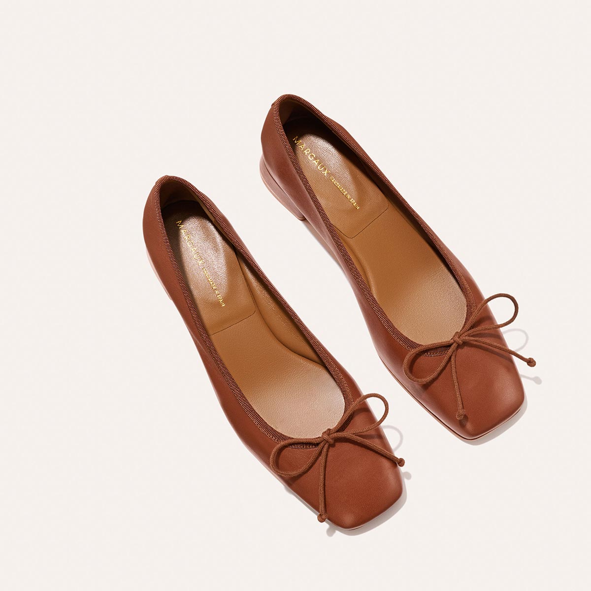 The Ada - Cognac Nappa by margaux-development: brown leather ballet flats with square toes and delicate bows, made using sacchetto construction for a glove-like fit, shown on a light background.
