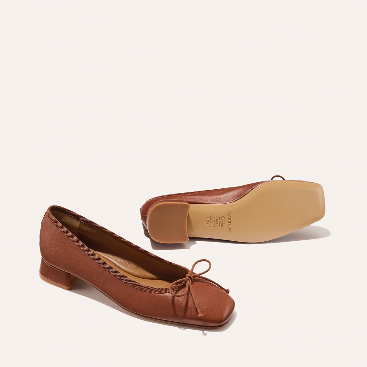 The Ada - Cognac Nappa by margaux-development: brown leather ballet pumps with low heels, square toes, bow detail, and a glove-like fit—one shoe stands upright while the other lies on its side to show the sole.