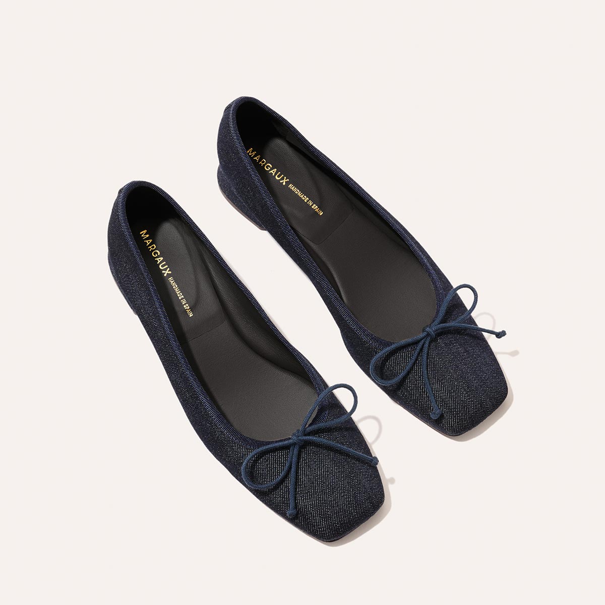 The Ada – Indigo Denim by margaux-development: A pair of indigo denim ballet flats with square toes and small bows on the front, shown on a light background. Black insoles with gold branding evoke classic style and elegance.