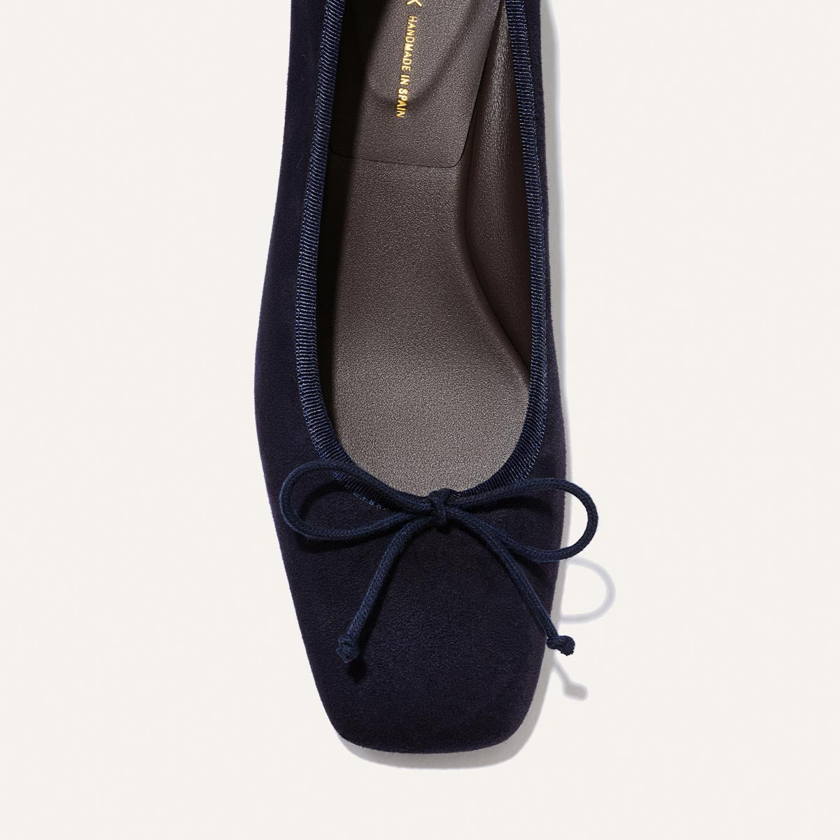 A close-up top view of The Ada - Indigo Suede by Margaux shows a dark navy blue low-heeled ballet pump with a square toe, small front bow, and light brown inner sole featuring partially visible gold branding.