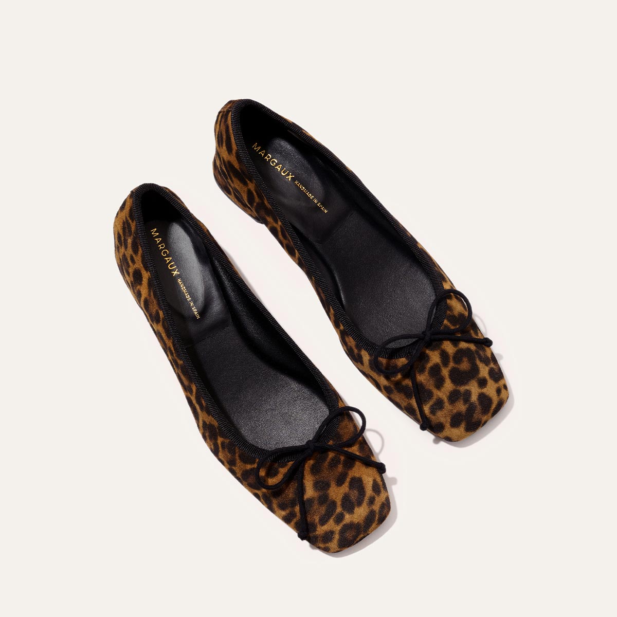 The Ada - Leopard Suede by margaux-development: leopard print ballet flats featuring a glove-like fit, black insoles, and small black bows on the toes, shown on a light background.