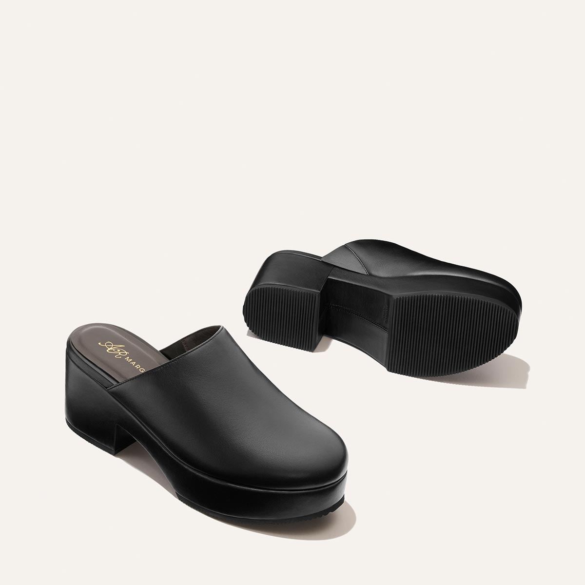 A pair of Margaux Alison Roman x Margaux: The Atlantic Clog in Black Calf—black leather slip-on clogs with chunky heels and ridged soles—are displayed on a light background. One shoe is upright, while the other rests on its side to reveal the sole.