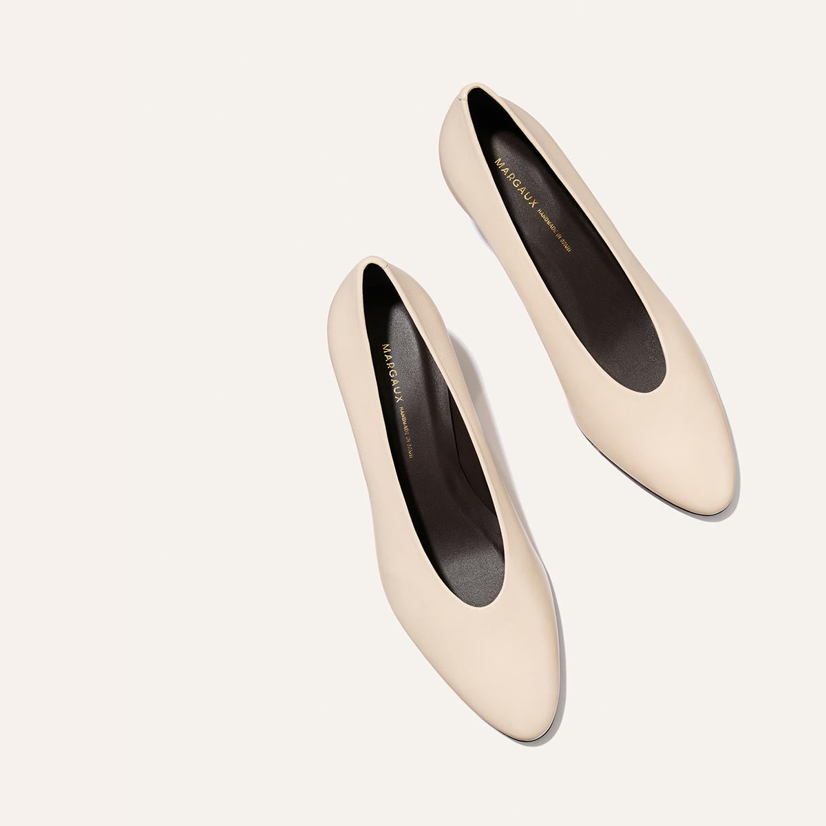 A pair of Margaux’s The Augusta Glove Pump flats in Bone Nappa, featuring a cream-colored, round-toe design and black insoles, is displayed diagonally on a plain, light background with the toes pointing toward the bottom right.