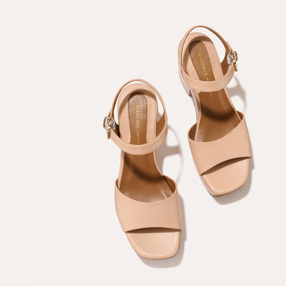 The Bardi - Petal Calf by margaux-development are nude, open-toe block heel sandals with ankle straps and gold buckles, bringing modern style to any look, shown on a light background.