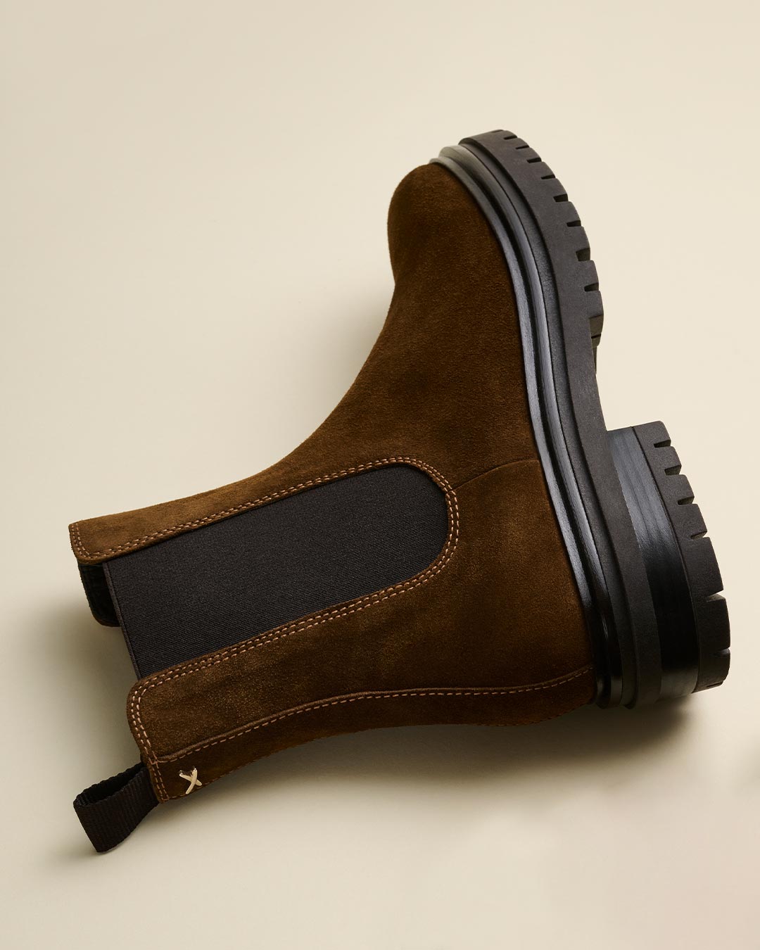 A brown suede Chelsea boot with black elastic side panels and a thick black rubber sole, displayed on its side against a light beige background.