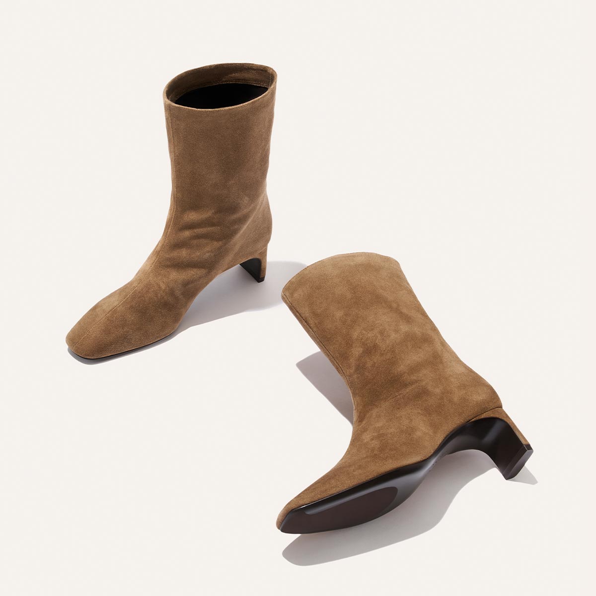 Two Brera Boots in Driftwood Suede by Margaux are positioned on a light background—one stands upright while the other lies on its side, displaying the sole and highlighting their mid-calf silhouette and subtle heel.