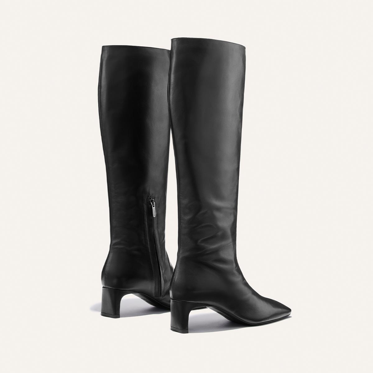 A pair of Margaux’s The Brera Knee-High Boot in Black Calf—tall black leather boots featuring mid-height block heels and side zippers—displayed on a plain light background.