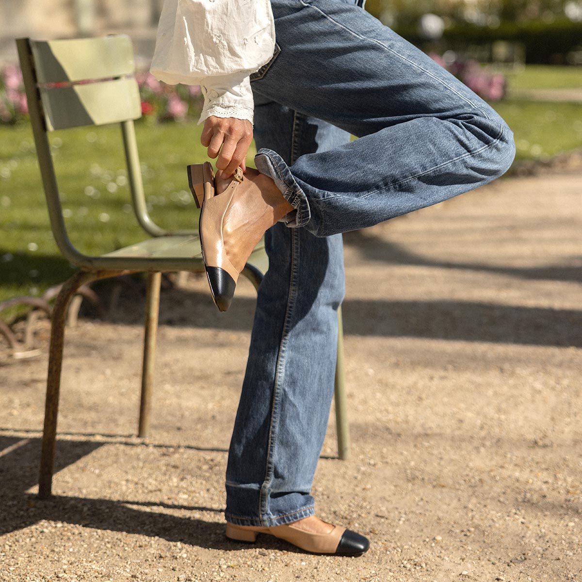 A person wearing blue jeans and Margauxs bestselling Darcy Slingback in Dune and Black Nappa balances on one leg by a green metal chair outdoors on a sunny, warm day.
