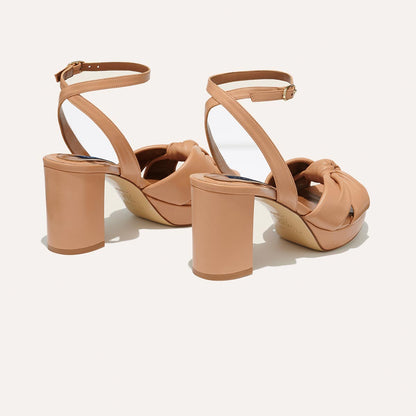The Carmine Platform - Desert Nappa by margaux-development features tan high-heeled sandals with ankle straps, chunky block heels, and a plush knot detail on the front, shown on a light background.