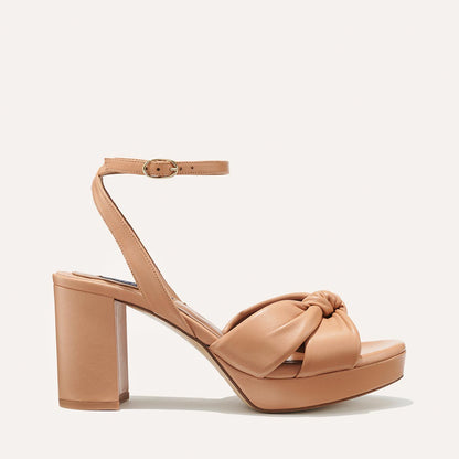 The Carmine Platform - Desert Nappa by margaux-development is a tan high-heeled sandal featuring a plush knot toe strap, thick platform sole, ankle buckle strap, and comfortable block heel, shown on a plain light background.