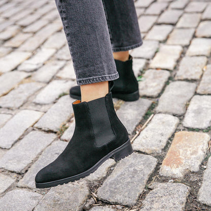 A person wearing Margaux’s The Chelsea Boot in black suede and cropped dark jeans walks on a cobblestone street.