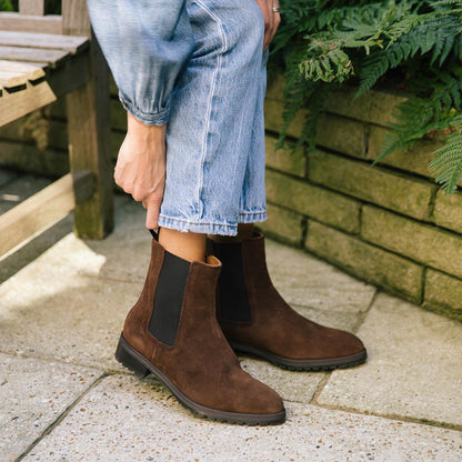 A person in cropped blue jeans adjusts The Chelsea Boot - Umber Suede by Margaux while standing outdoors on a stone patio beside a brick planter of green ferns.