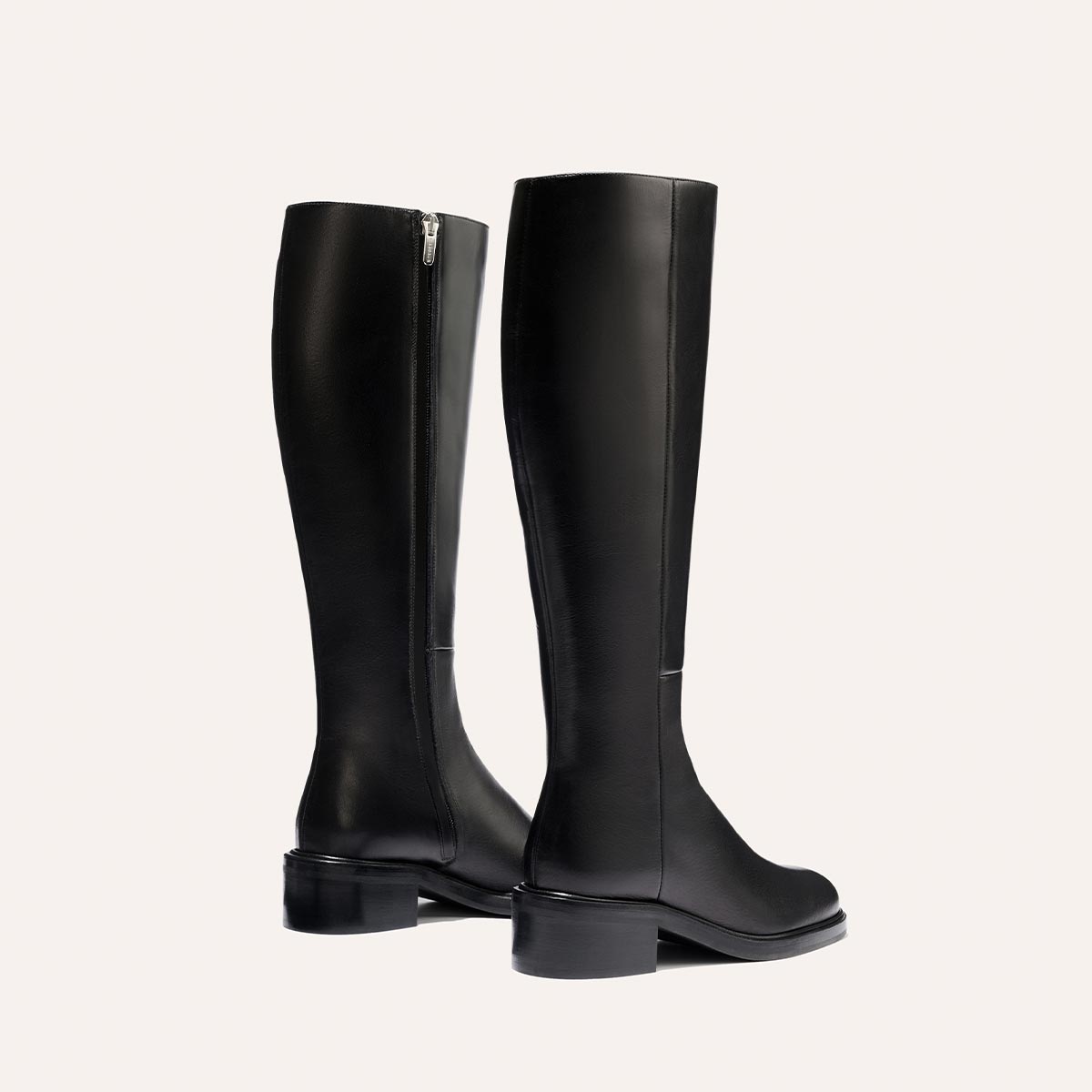 The City Boot - Black Calf by Margaux: a pair of tall black leather boots with a classic silhouette, side zippers, and low block heels, shown on a light background—one boot faces forward, the other turned slightly to the side.