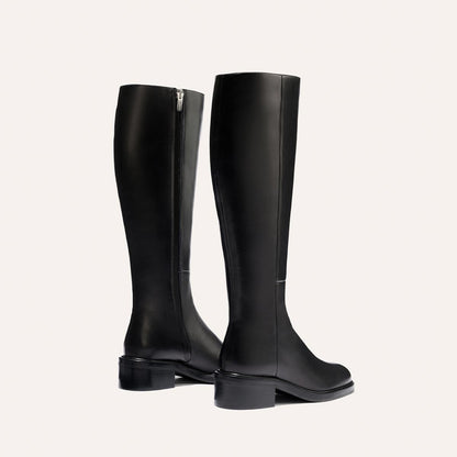 The City Boot - Black Calf by Margaux: a pair of tall black leather boots with a classic silhouette, side zippers, and low block heels, shown on a light background—one boot faces forward, the other turned slightly to the side.