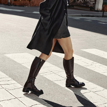 A person wearing Margaux's The City Boot - Black Calf walks across a crosswalk on a sunny day, paired with a black coat and gray skirt.