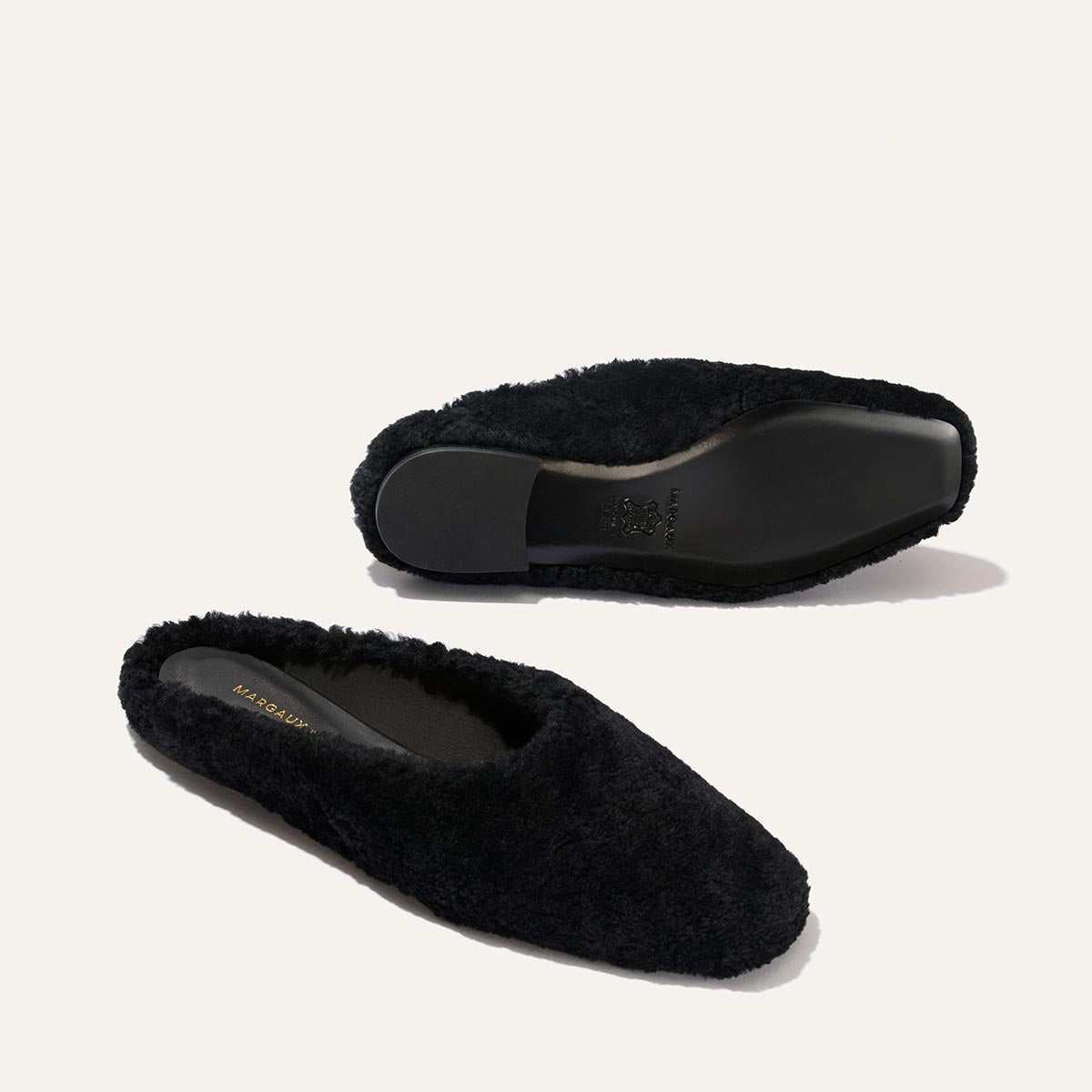 Two black shearling slippers, The Clara by Margaux, are displayed on a light background. One slipper stands upright, showcasing the do-it-all style and soft upper, while the other is upside down to reveal its flat black sole and versatile slipper-like silhouette.
