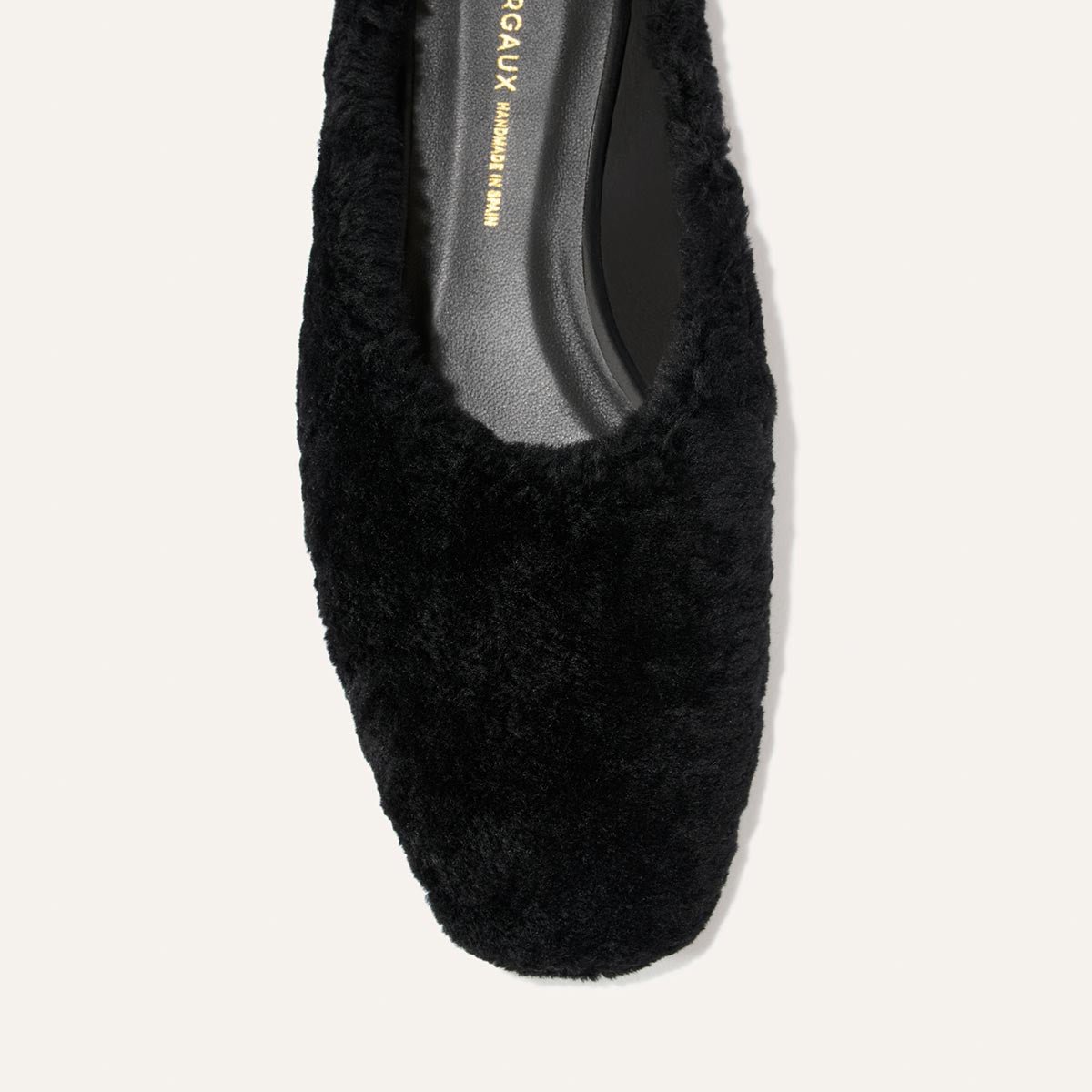 A close-up of The Clara - Black Shearling by Margaux, a black flat shoe featuring plush shearling texture, a square toe, and a black insole, shown from above against a light background. The slipper-like silhouette enhances the versatile appeal of this do-it-all style.