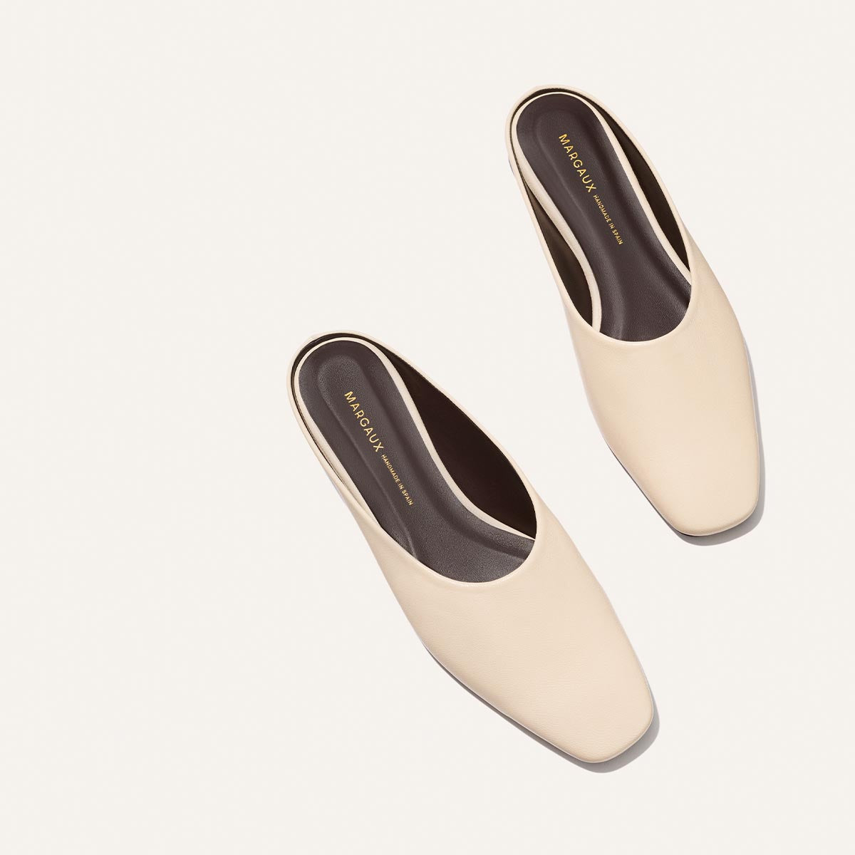 A pair of The Clara - Bone Nappa backless slip-on shoes by Margaux with square toes, shown from above on a light background. The dark insoles feature "Margaux" and "New York" printed in gold lettering.