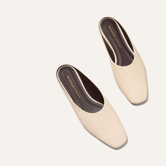 A pair of The Clara - Bone Nappa backless slip-on shoes by Margaux with square toes, shown from above on a light background. The dark insoles feature "Margaux" and "New York" printed in gold lettering.