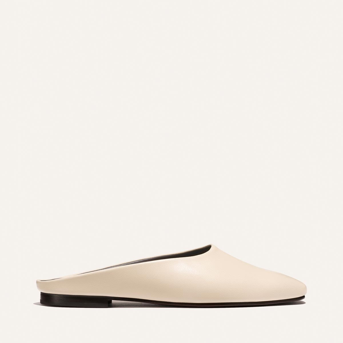 The Clara - Bone Nappa by Margaux is a minimalist, cream-colored slip-on mule with a closed toe and flat black sole, shown from the side against a plain white background.