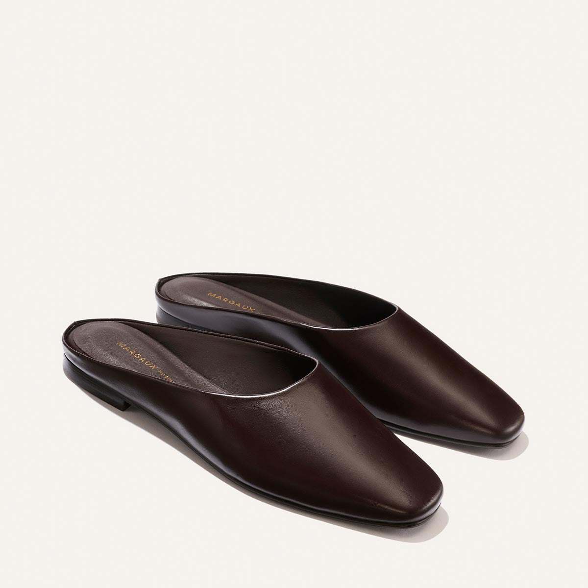 Introducing The Clara - Coffee Calf by Margaux: a pair of dark brown leather slip-on mules with a flat, versatile design and closed, rounded toes. Featuring a slipper-like silhouette, these shoes are beautifully displayed on a light background.
