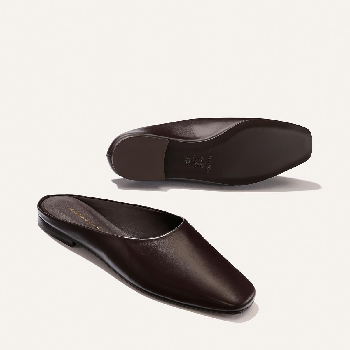 The Clara - Coffee Calf by Margaux, a pair of dark brown leather slip-on mule shoes, is showcased on a light beige background with one shoe upright and the other displaying the sole. Boasting a slipper-like silhouette, these flats embody simple, elegant minimalism for effortless, do-it-all style.