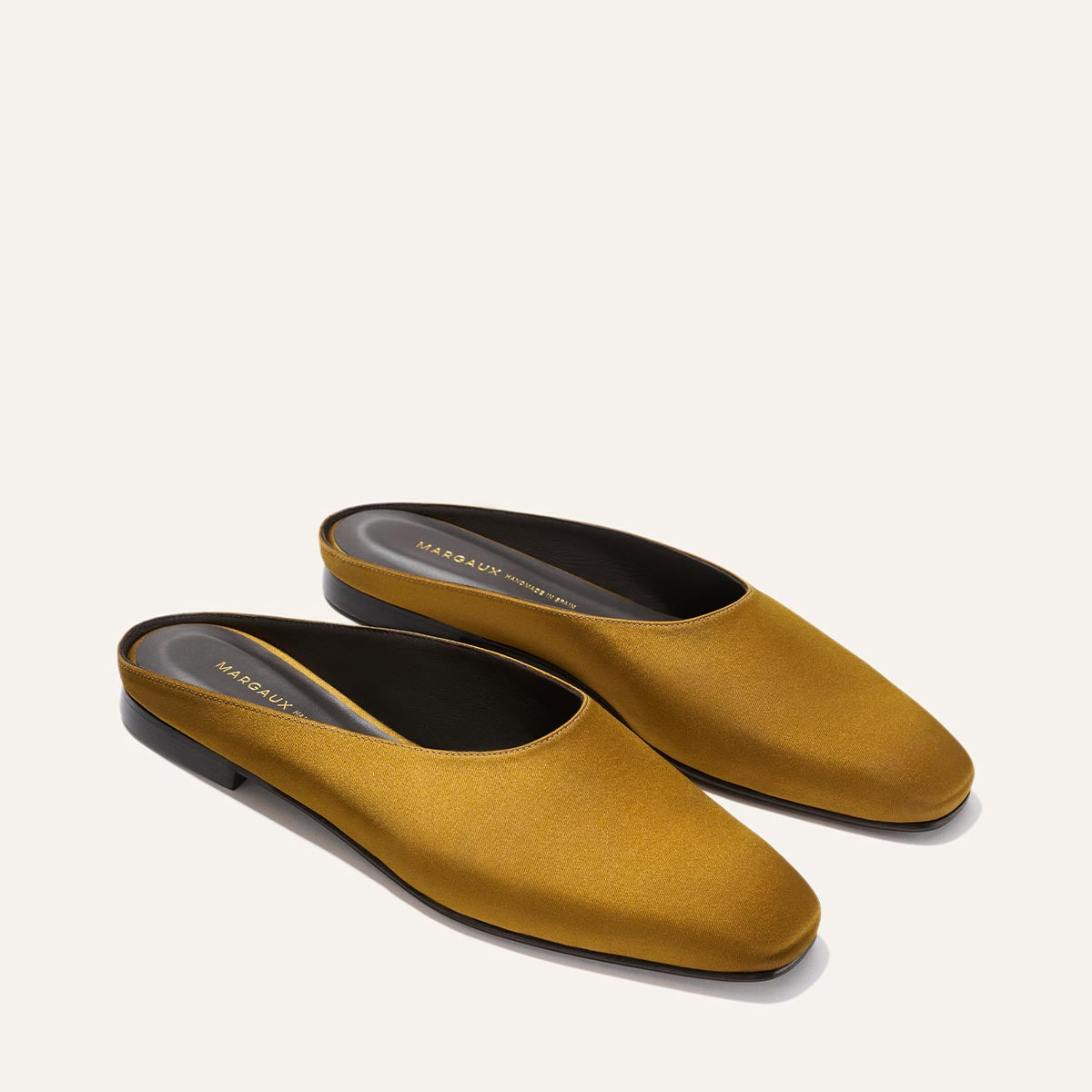 The Clara - Ochre Satin by Margaux: mustard yellow slip-on mules featuring a low black heel and rounded square toe, shown on an off-white background.