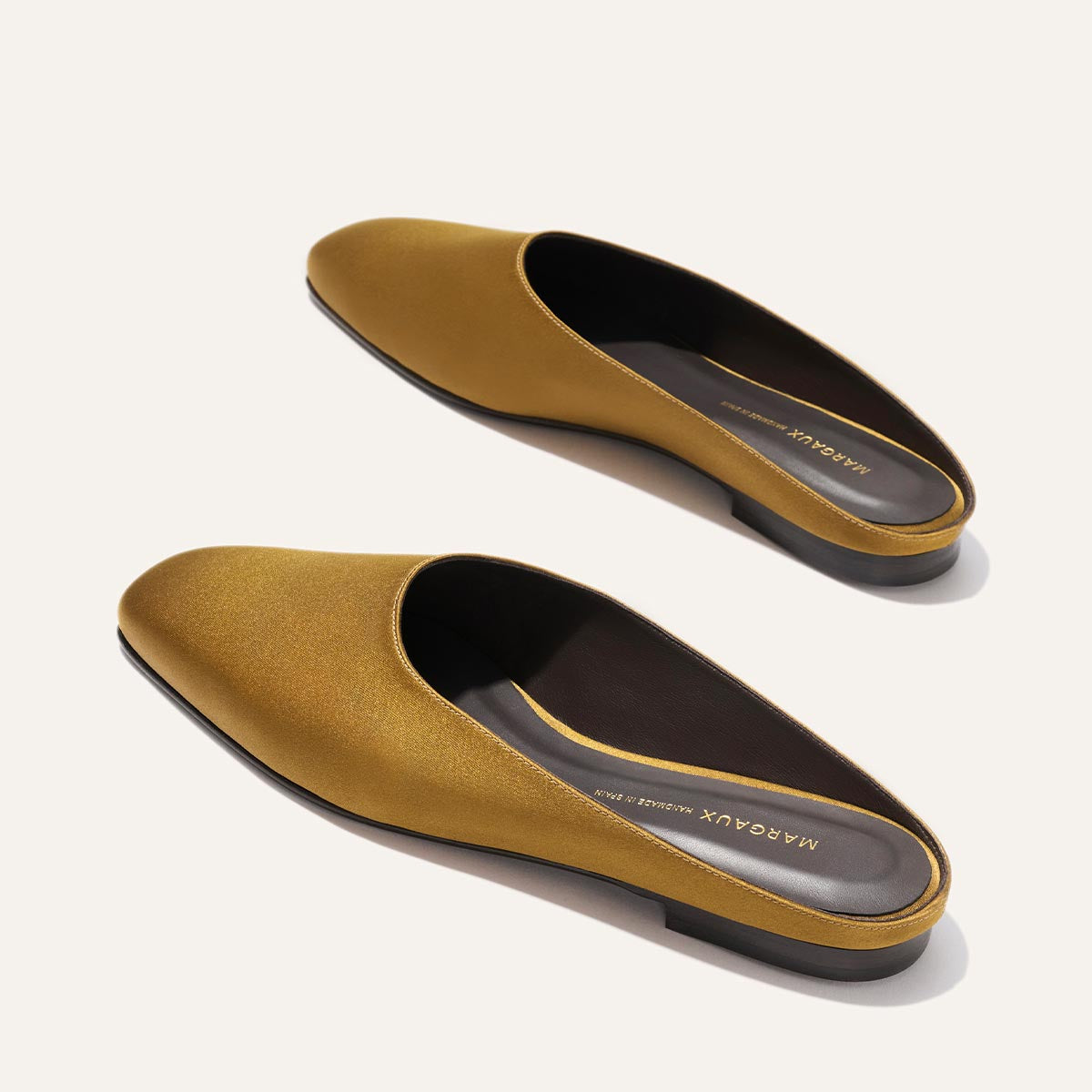 The Clara - Ochre Satin by Margaux are mustard yellow slip-on mules with a low, flat heel and sleek, minimalist design, photographed on a light background.