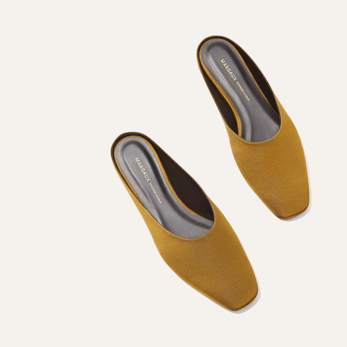 A pair of The Clara - Ochre Satin mules by Margaux, featuring a square toe and slip-on design, are displayed diagonally on a light beige background. The gray insole is stamped with “MARGAUX” in gold lettering.