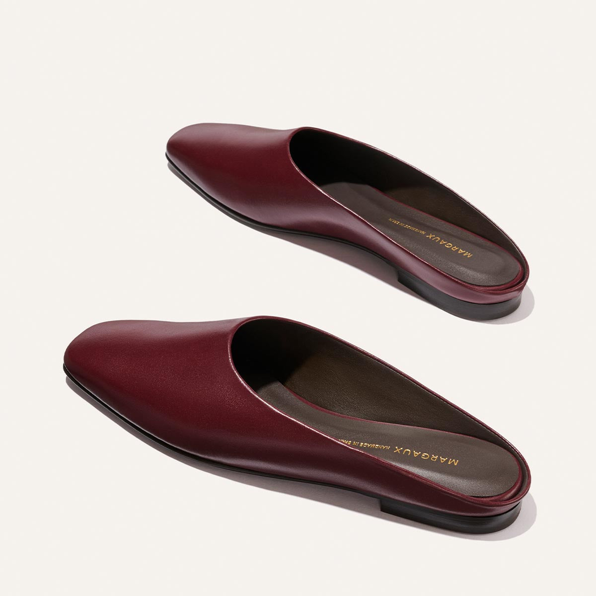 Introducing The Clara - Wine Calf by Margaux: a pair of versatile burgundy leather slip-on mules featuring square toes and low black heels in a classic slipper silhouette, elegantly displayed on a light background.