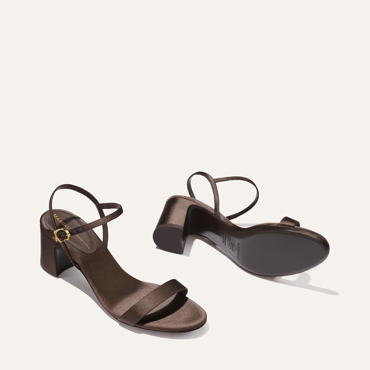 The Cleo Sandal - Espresso Satin by Margaux: a brown, open-toe sandal with a low block heel and slim ankle straps with gold buckles. One stands upright, the other lies on its side to display the sole.