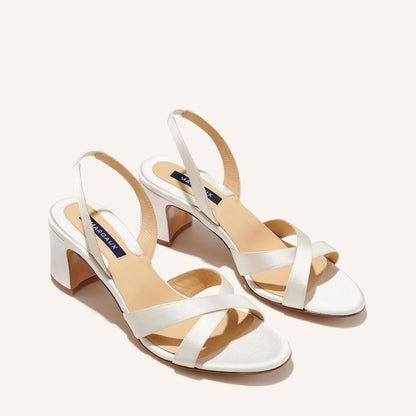 The Clotilde Sandal in Ivory Satin by margaux-development features block heels, crisscross front straps, and slingback ankle straps, shown on a light background.