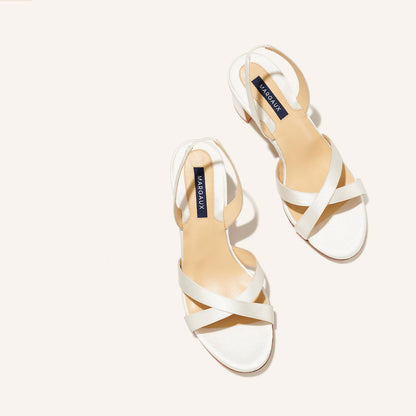 The Clotilde Sandal - Ivory Satin by margaux-development features white open-toe sandals with crisscross straps, low heels, and tan insoles with a black margaux-development label, displayed on a light background.