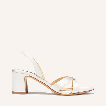 The Clotilde Sandal - Ivory Satin by margaux-development features white satin crisscross straps, a slingback heel strap, an open toe, and a mid-height block heel for a minimalist, elegant look with a light tan sole.