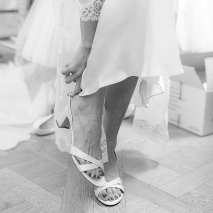 A person in a white dress, shown from the knees down, adjusts The Clotilde Sandal - Ivory Satin by margaux-development on their foot. The black and white image features a wooden floor and a white box in the background.