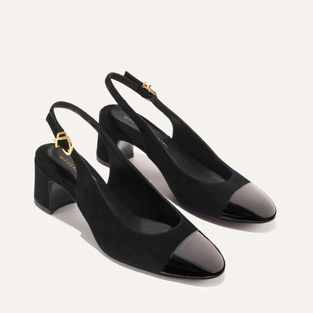 The Cluny Slingback by Margaux features black suede uppers, shiny patent closed toes, gold buckles, and low block heels—an elegant work shoe—displayed against a light background.