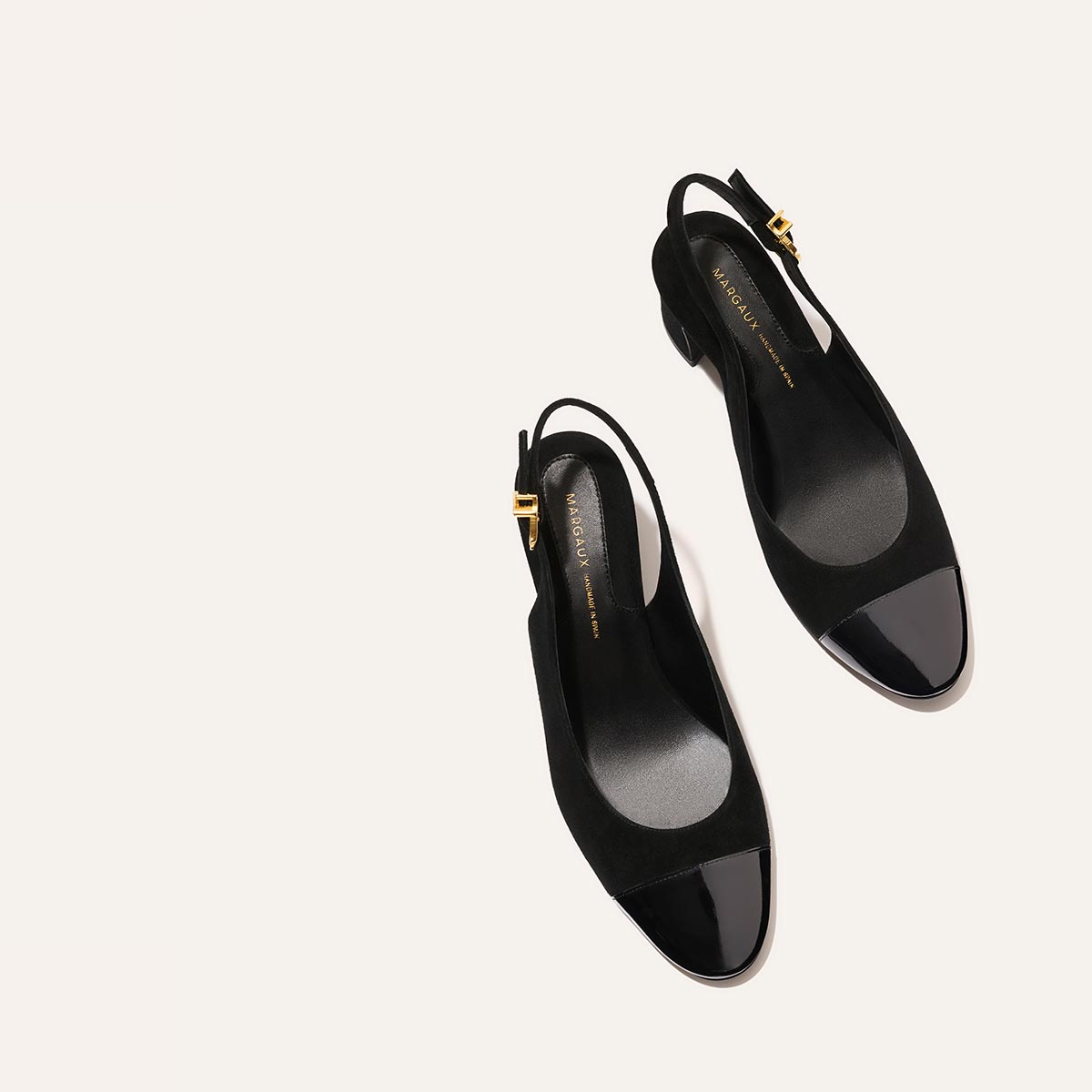 The Margaux Cluny Slingback in black suede and patent features shiny black toe caps, low heels, and gold buckle accents—elegant and versatile heels for work or special occasions, shown on a light beige background.
