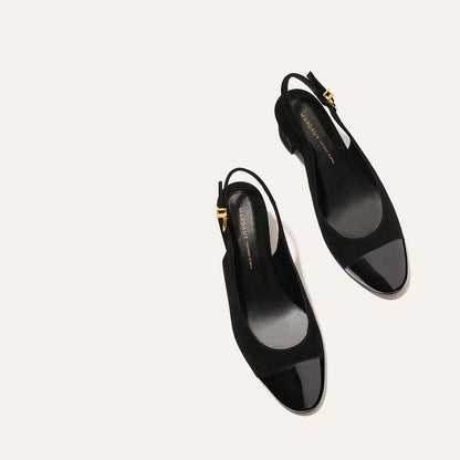 The Margaux Cluny Slingback in black suede and patent features shiny black toe caps, low heels, and gold buckle accents—elegant and versatile heels for work or special occasions, shown on a light beige background.