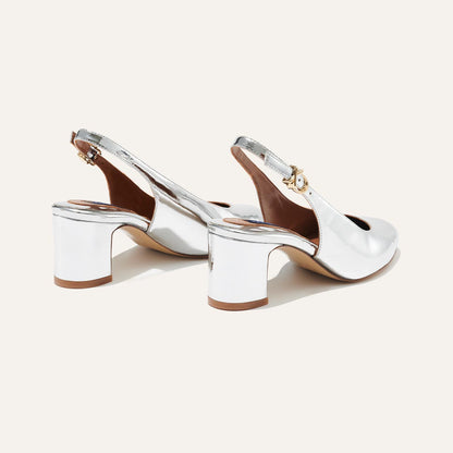 The Cluny Slingback - Silver Mirror by Margaux features shiny silver block heels and ankle straps, exuding Parisian elegance. Displayed on a plain light background, these pumps elevate any look with their refined details.