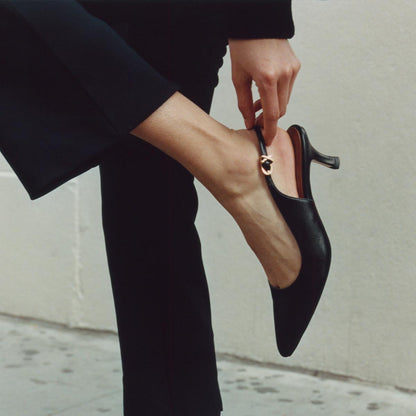 A person in black pants adjusts The Colette - Black Nappa snip nose slingback with a micro heel from margaux-development, their hand on the strap. The background shows a light-colored wall and floor.